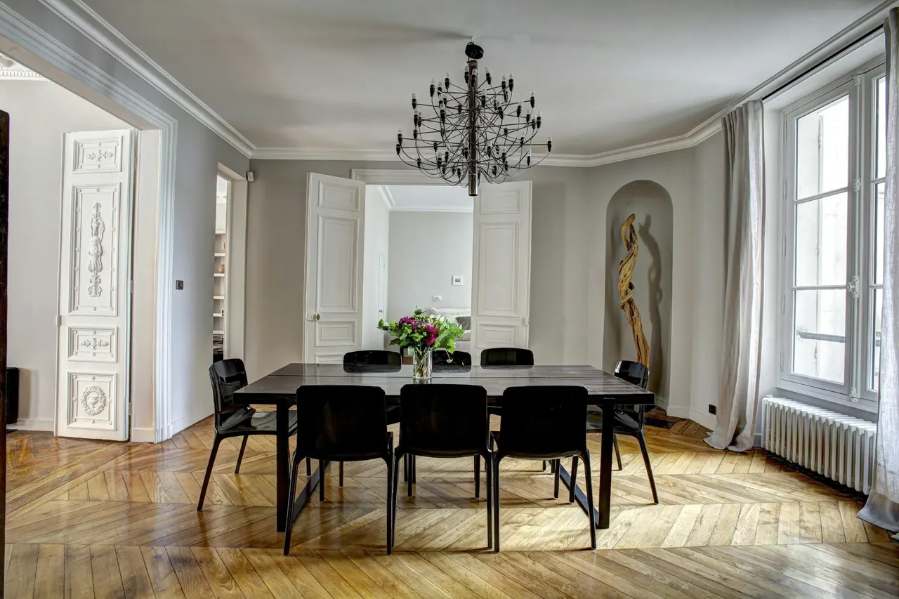 Comedor con mesa larga, sillas negras, lámpara de araña y suelo de espiga